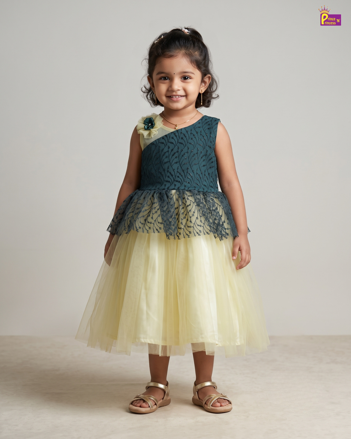 A young girl stands against a plain light gray studio background wearing a sleeveless party dress with a dark teal lace bodice, a pale yellow tulle skirt, and a small floral embellishment on one shoulder. She is also wearing metallic sandals.