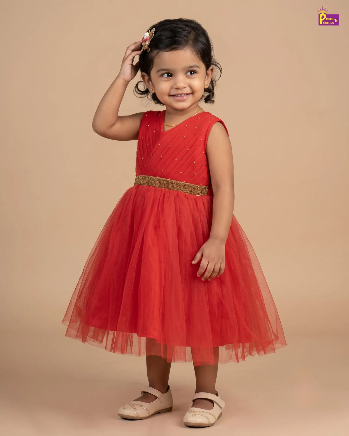 A young girl modeling a sleeveless red party dress with a gold metallic waistband and a full tulle skirt, standing against a solid beige studio background.