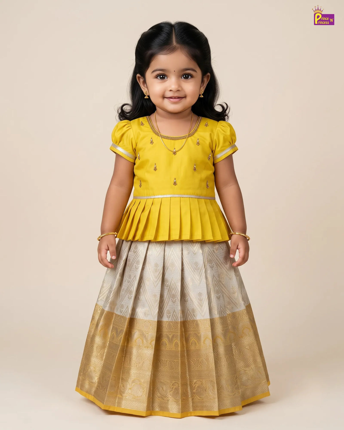 A young girl modeling a traditional Indian outfit consisting of a bright yellow peplum top with silver embroidery and a matching flared skirt in beige and gold with a brocade border, set against a solid beige background.