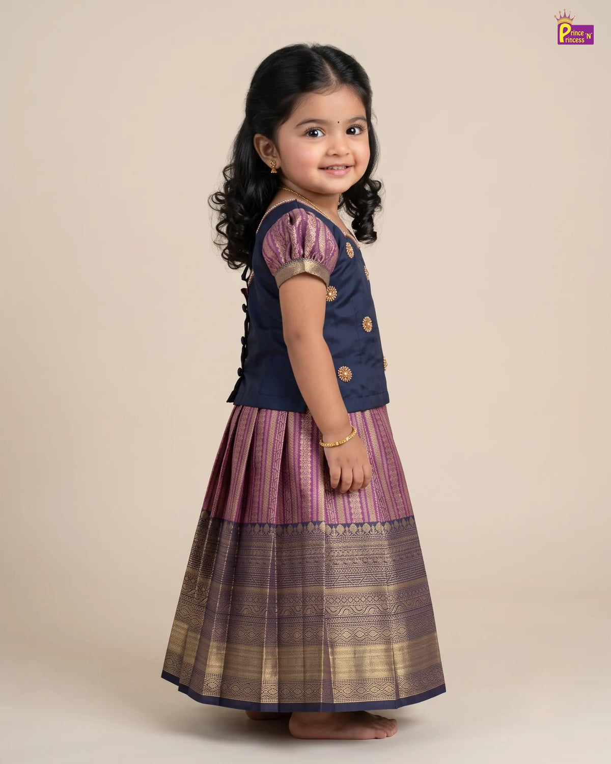 A young girl modeling a traditional Indian outfit consisting of a navy blue sleeveless top with gold buttons and a pleated purple and gold skirt, set against a solid beige background.