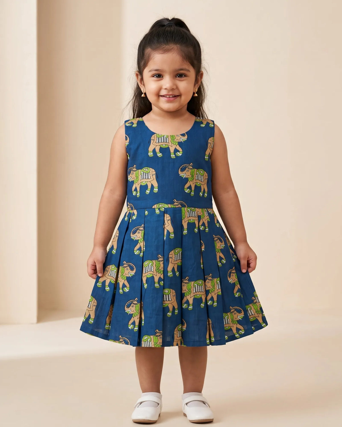 A young girl modeling a traditional Indian dress consisting of an olive green sleeveless kurta and a matching pleated skirt with gold and maroon borders, set against a plain beige studio background.