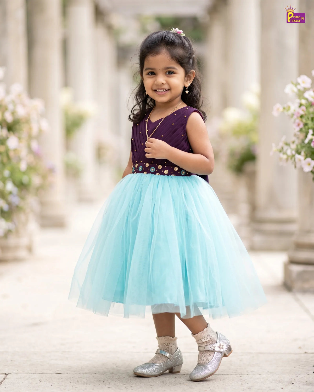 A young girl stands in a sunlit outdoor corridor wearing a sleeveless party dress with a deep purple bodice, floral embroidery, and a voluminous light aqua tulle skirt. She is wearing sparkly silver shoes and socks.