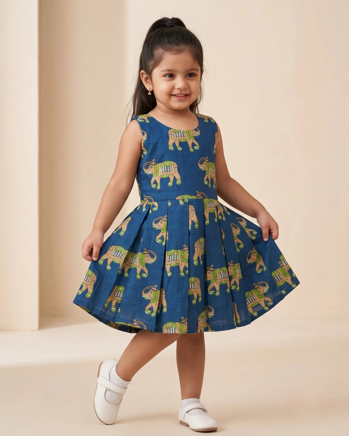 A young girl with her hair in a ponytail is wearing a sleeveless blue dress with a pattern of elephants carrying yellow and green loads. She is standing in a studio with a beige background, wearing white shoes.