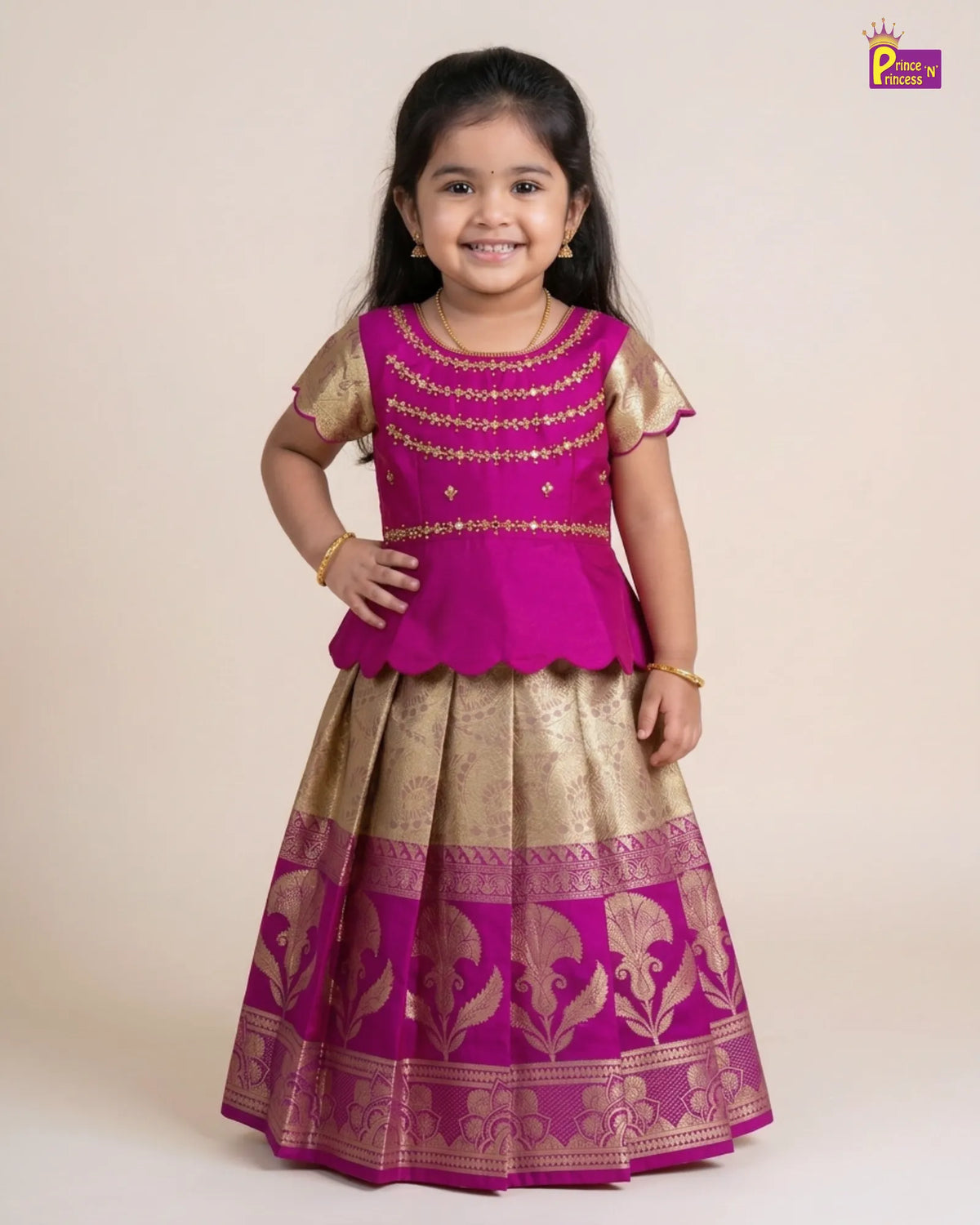 A young girl modeling a traditional Indian outfit consisting of a bright magenta pink kurta with gold embroidery and a matching pleated skirt with gold zari patterns, set against a plain beige studio background.