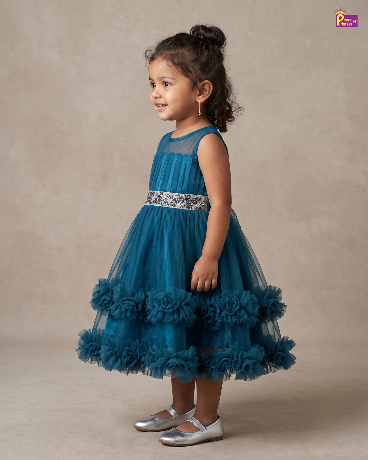 A young girl with her hair in a high bun stands in profile wearing a sleeveless teal blue tulle dress with a floral rosette skirt and a silver patterned waistband, paired with silver shoes, against a neutral beige studio backdrop.