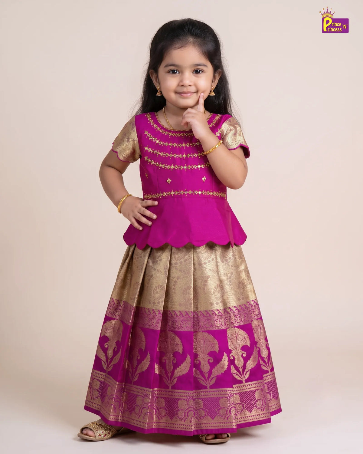 A young girl modeling a traditional Indian outfit consisting of a bright magenta peplum top and a matching pleated skirt with gold zari embroidery, set against a plain beige studio background.