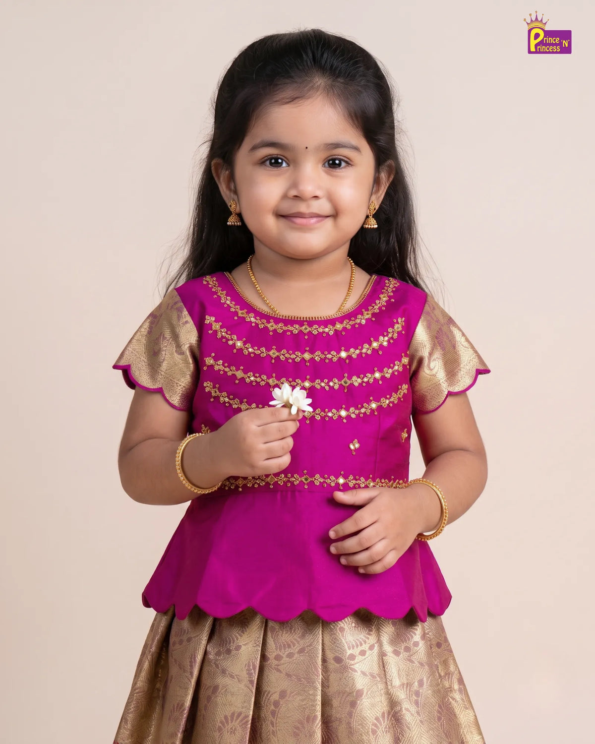 A young girl modeling a traditional Indian outfit consisting of a bright magenta pink top with gold embroidery and a matching gold patterned skirt, set against a plain beige background.