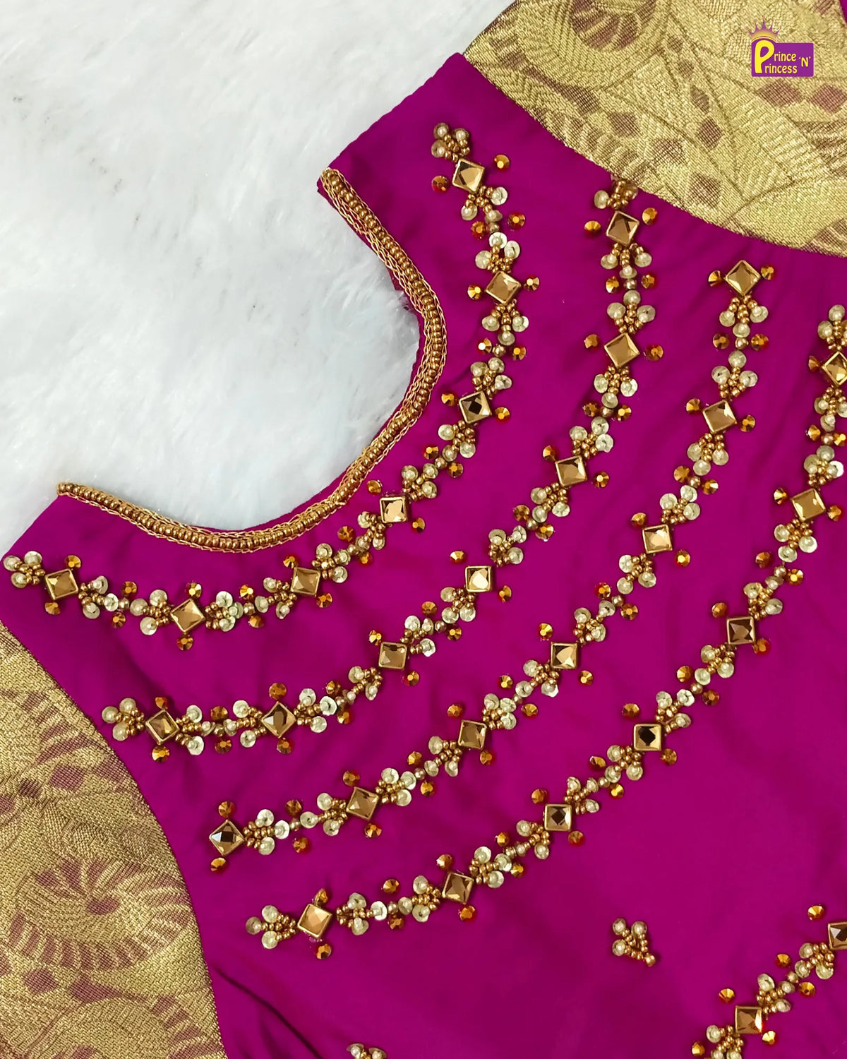A close-up of a bright magenta pink fabric garment, likely a saree blouse, decorated with intricate gold zari embroidery and mirror work. The blouse features a scalloped neckline with gold border trim and detailed gold beadwork.