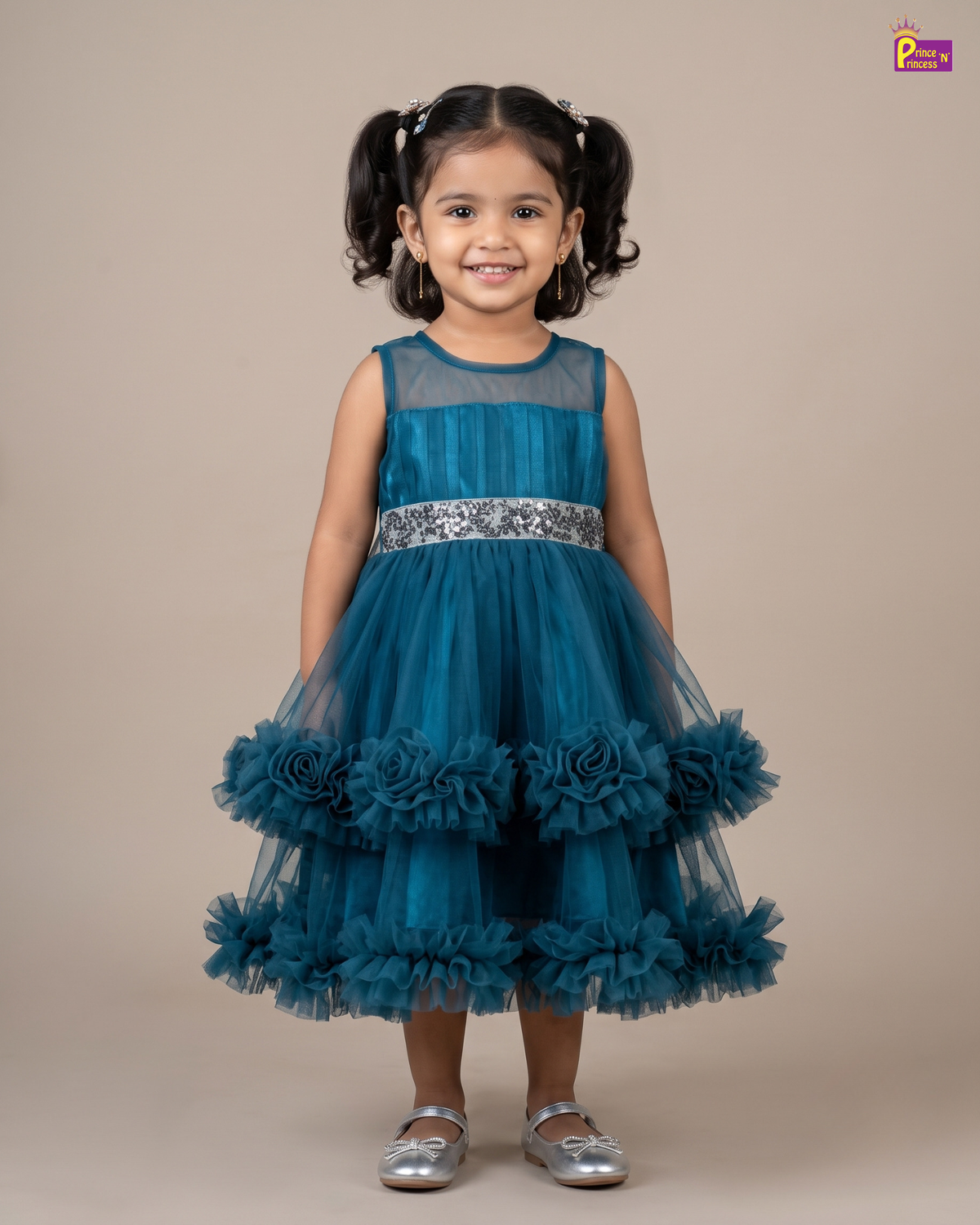 A young girl with dark hair in pigtails stands against a solid beige studio background wearing a sleeveless teal blue tulle dress with a silver patterned waistband and layered ruffle details on the skirt, paired with metallic silver shoes.
