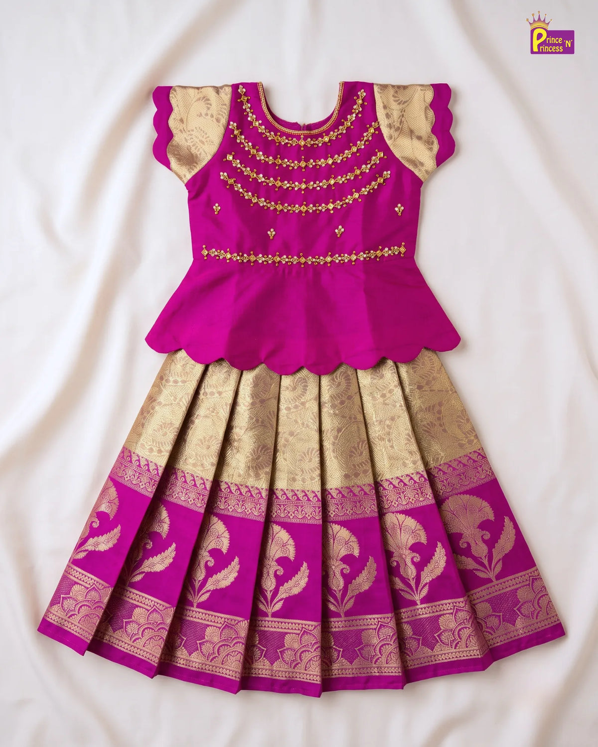 A traditional Indian girl's dress featuring a bright magenta pink peplum top with gold embroidery and a matching pleated skirt with gold zari patterns, laid flat on a white fabric background.