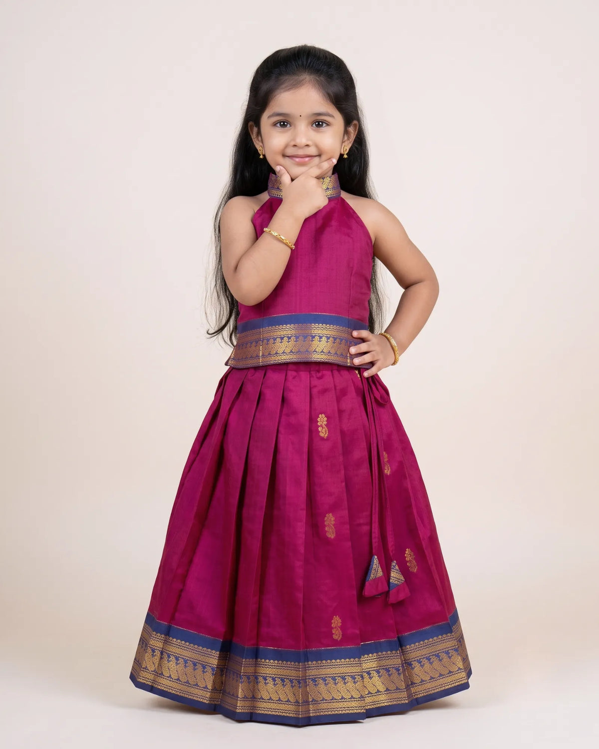 A young girl modeling a magenta pink traditional Indian dress with gold and blue zari borders, standing against a plain beige studio background.