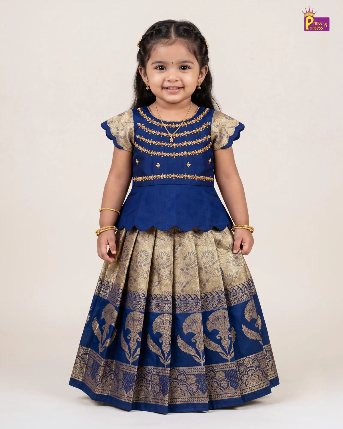 A young girl modeling a royal blue and gold traditional Indian ethnic dress with a peplum waist, gold embroidered bodice, and a heavily patterned gold skirt, set against a plain beige studio background.