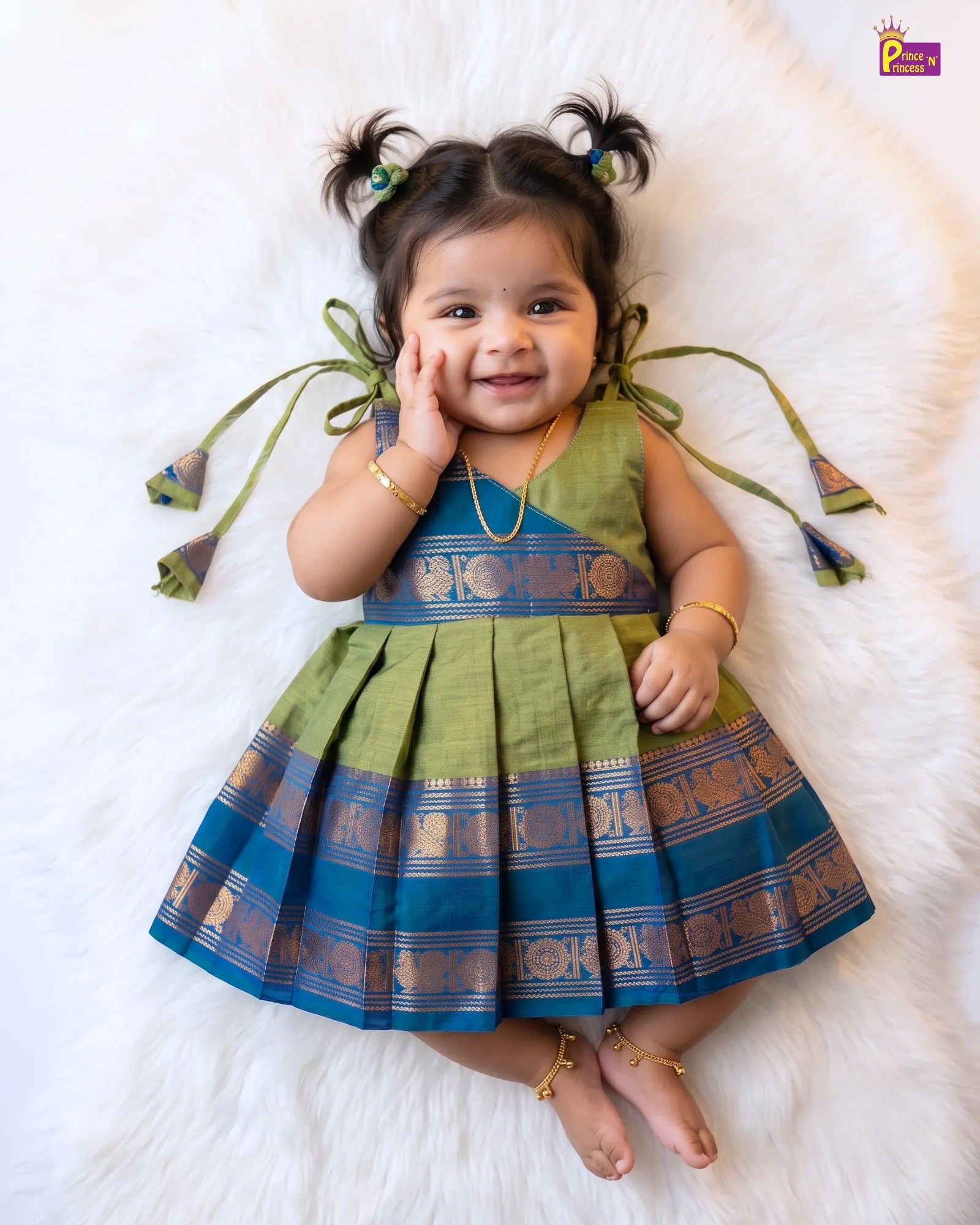A toddler girl lying on a white faux fur rug wearing a green and blue saree-style dress with gold zari patterns, gold jewelry, and her hair styled in two buns.