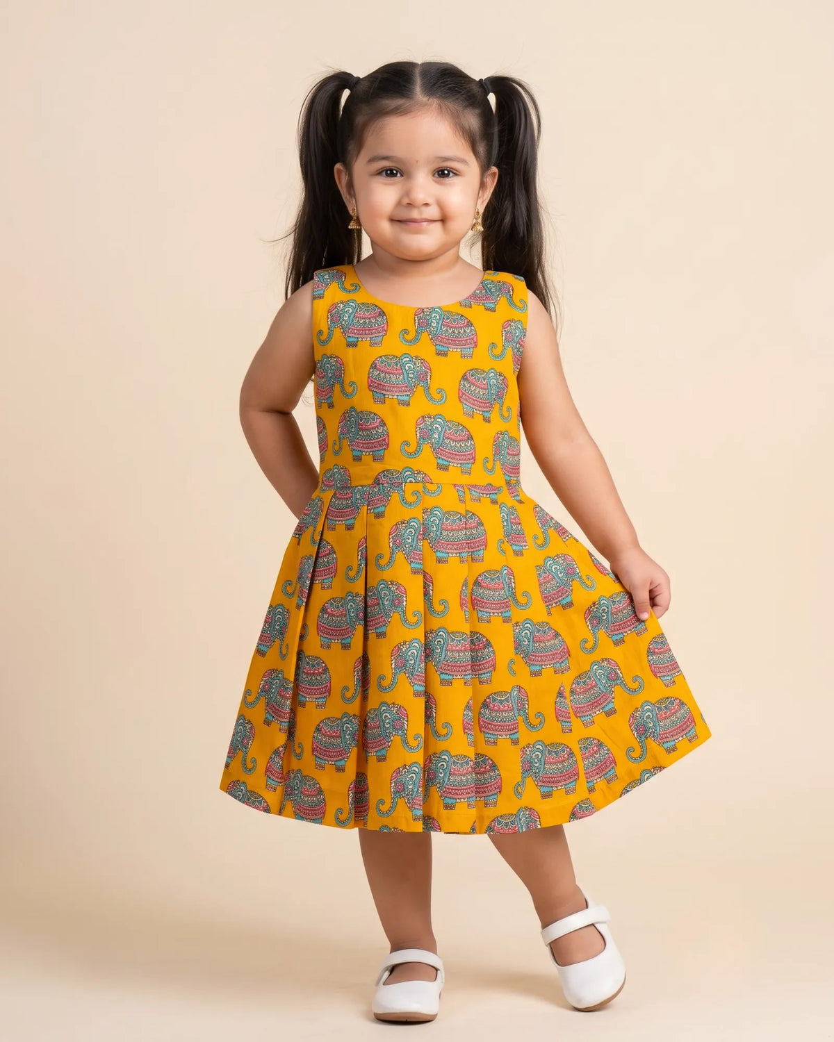 A young girl with long brown hair stands against a light beige studio background wearing a traditional maroon sleeveless dress with gold and green borders. She is posing with her hands on her cheeks.