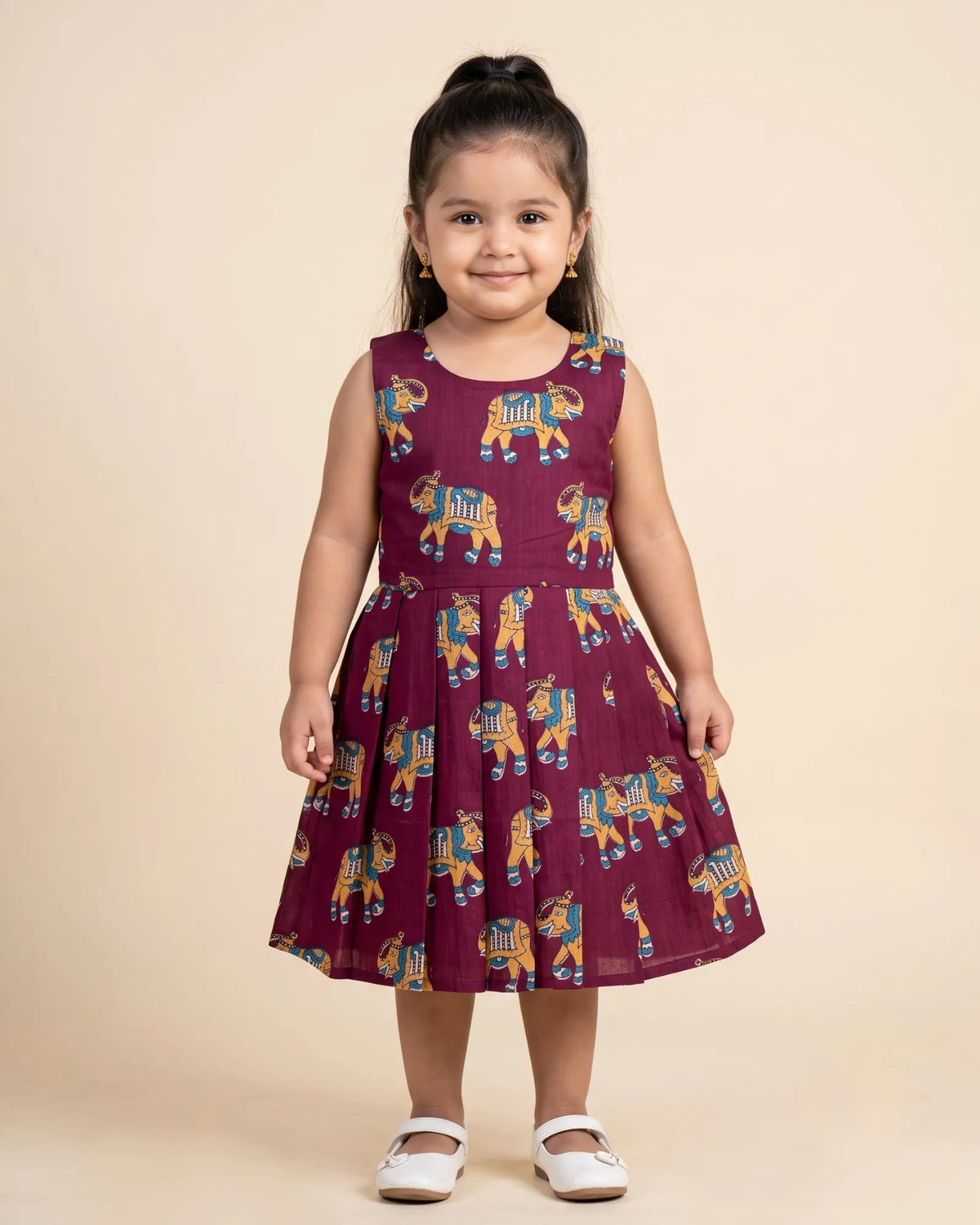 A young girl modeling a sleeveless maroon traditional dress with gold and black borders, standing against a plain light beige background.