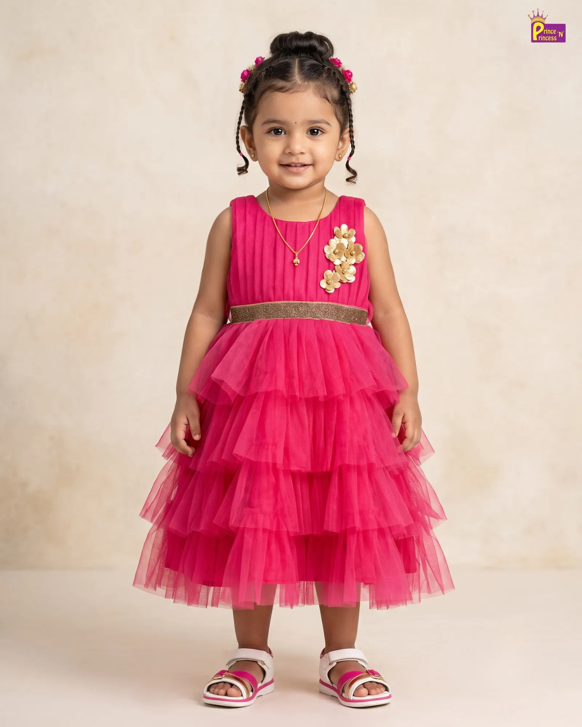 A young girl with braided hair and a flower crown is wearing a bright pink tiered tulle dress with a gold sequin waistband and a gold floral applique on the chest. She is standing against a neutral beige background.