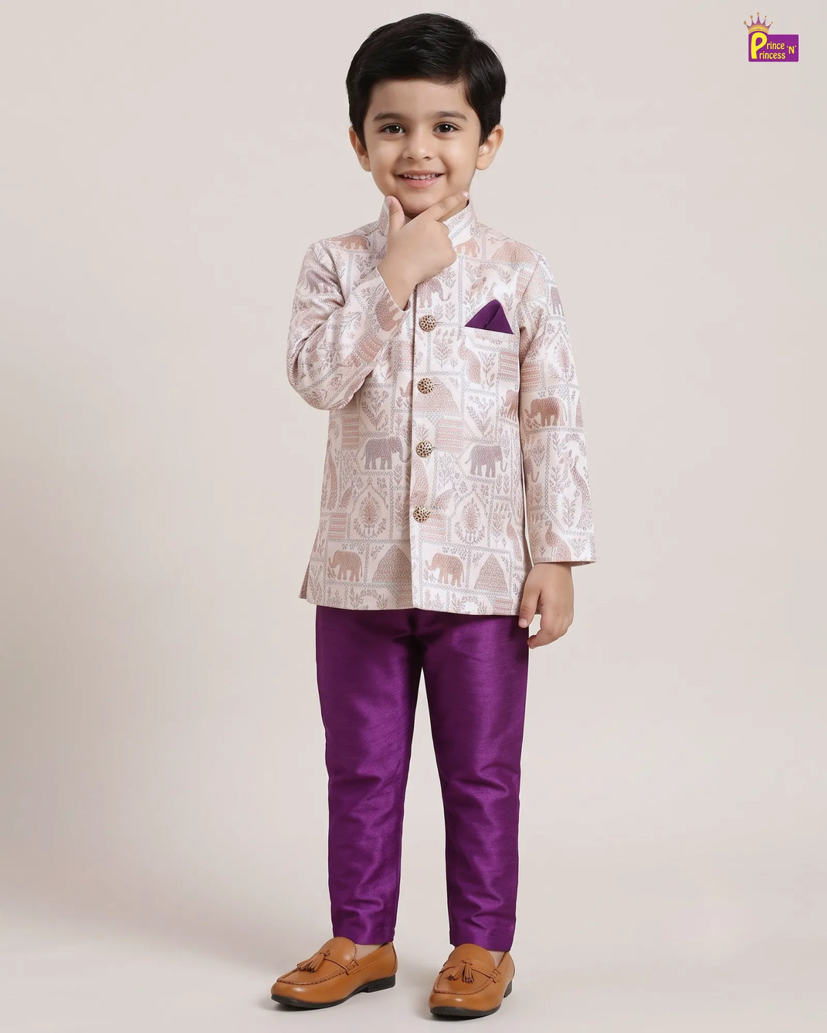 A young boy modeling a traditional Indian outfit consisting of a light beige printed kurta with a purple pocket square, paired with shiny purple trousers and brown tassel loafers, against a plain beige studio background.