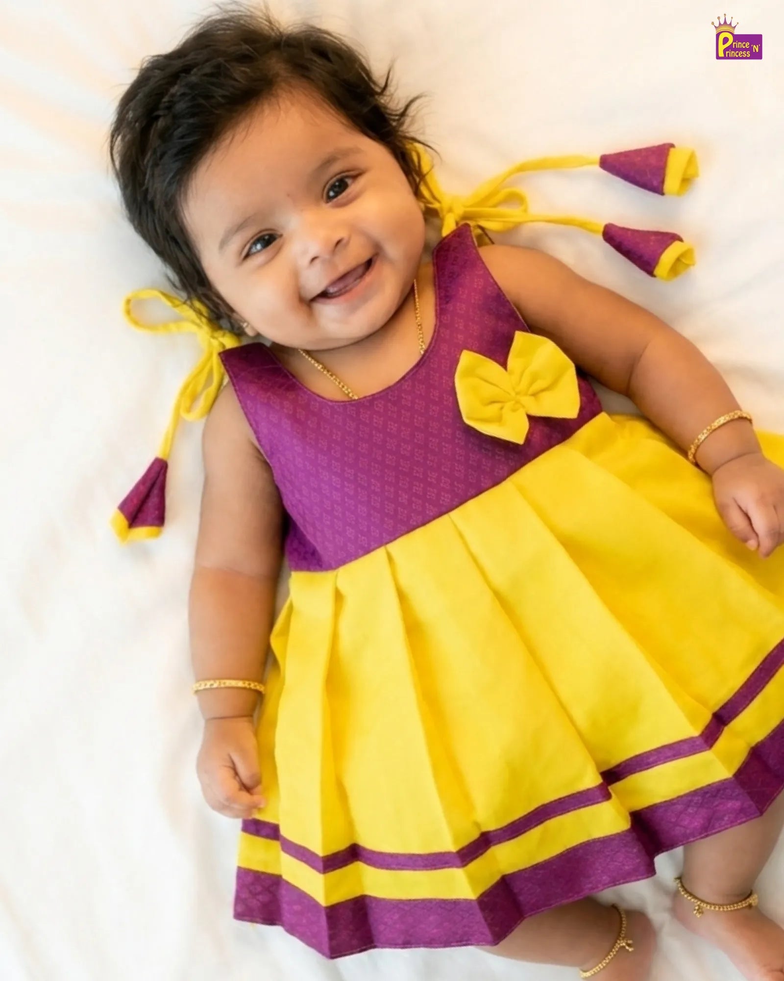 Child wearing a yellow and purple dress with a bow, lying on a white surface.