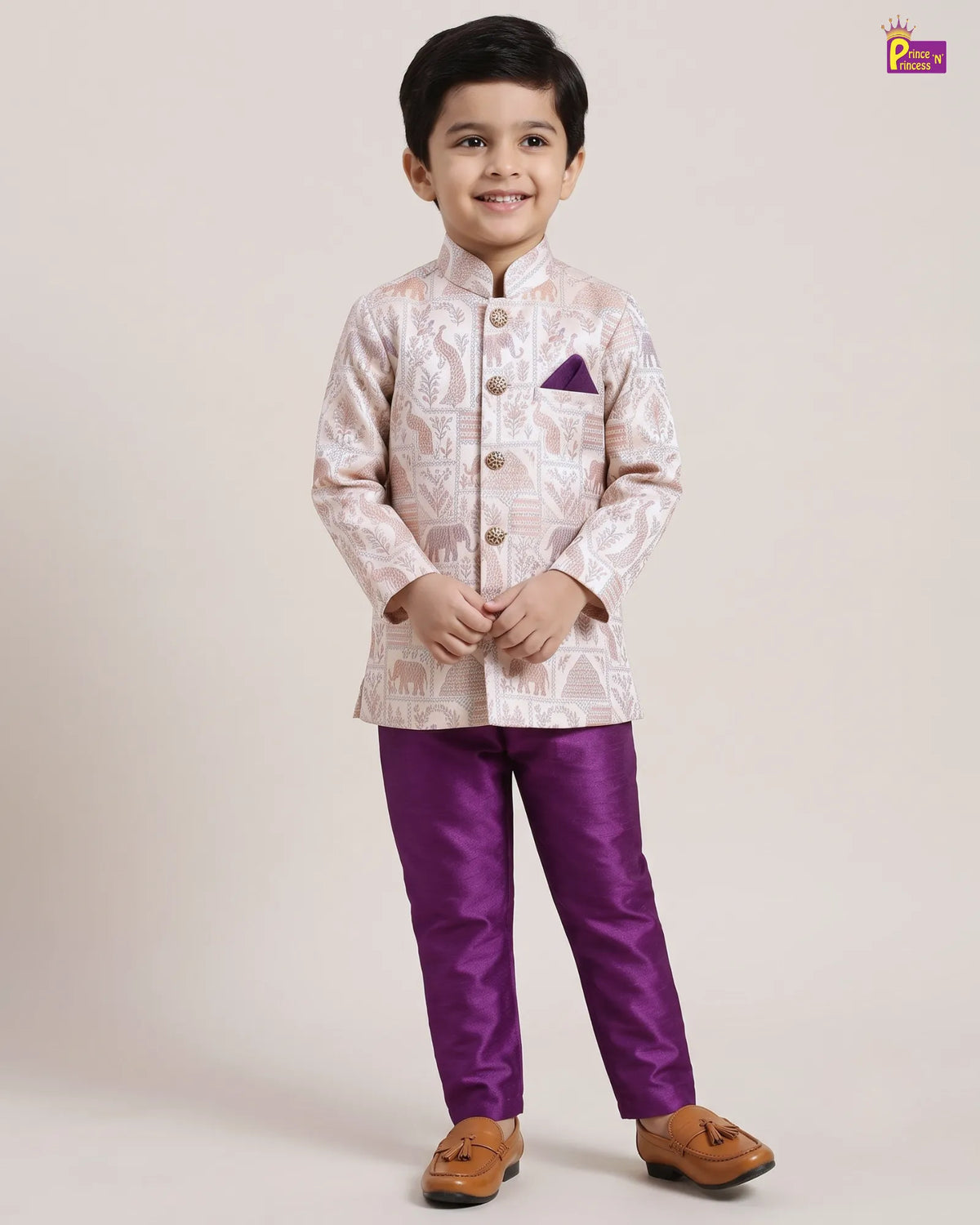 A young boy modeling a traditional Indian outfit consisting of a light beige and white patterned sherwani jacket with a purple pocket square, paired with shiny purple trousers and brown shoes, against a plain beige background.