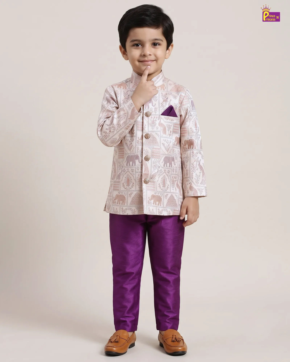 A young boy modeling a traditional Indian outfit consisting of a light beige printed sherwani jacket with elephant motifs and a purple pocket square, paired with solid purple trousers and brown tassel shoes, against a plain beige studio background.