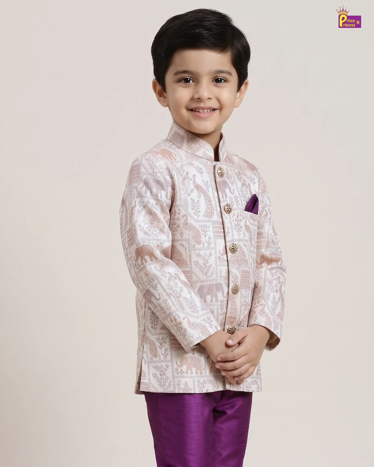 A young boy modeling a traditional Indian outfit consisting of a light pink and white patterned sherwani jacket with a purple pocket square and matching purple trousers, set against a plain beige studio background.