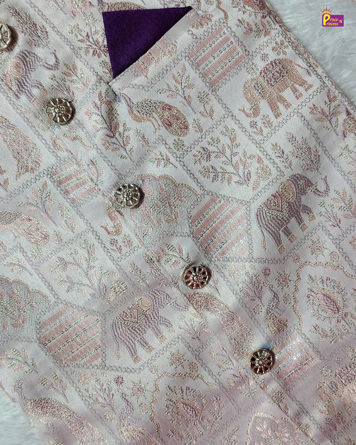 A close-up of a light pink and white brocade fabric with detailed elephant and floral motifs, adorned with metallic buttons and a purple folded corner.