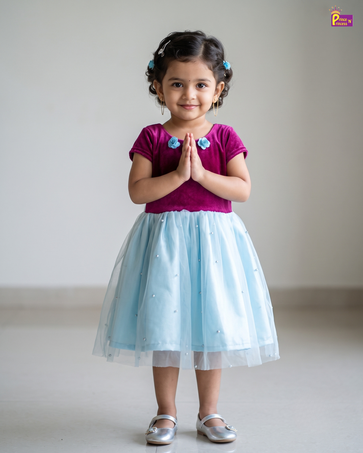 A young girl with her hair in two buns stands with her hands pressed together, wearing a magenta short-sleeve top with blue floral embroidery and a light blue tulle skirt with small embroidered details. She is wearing silver shoes against a plain light gray background.