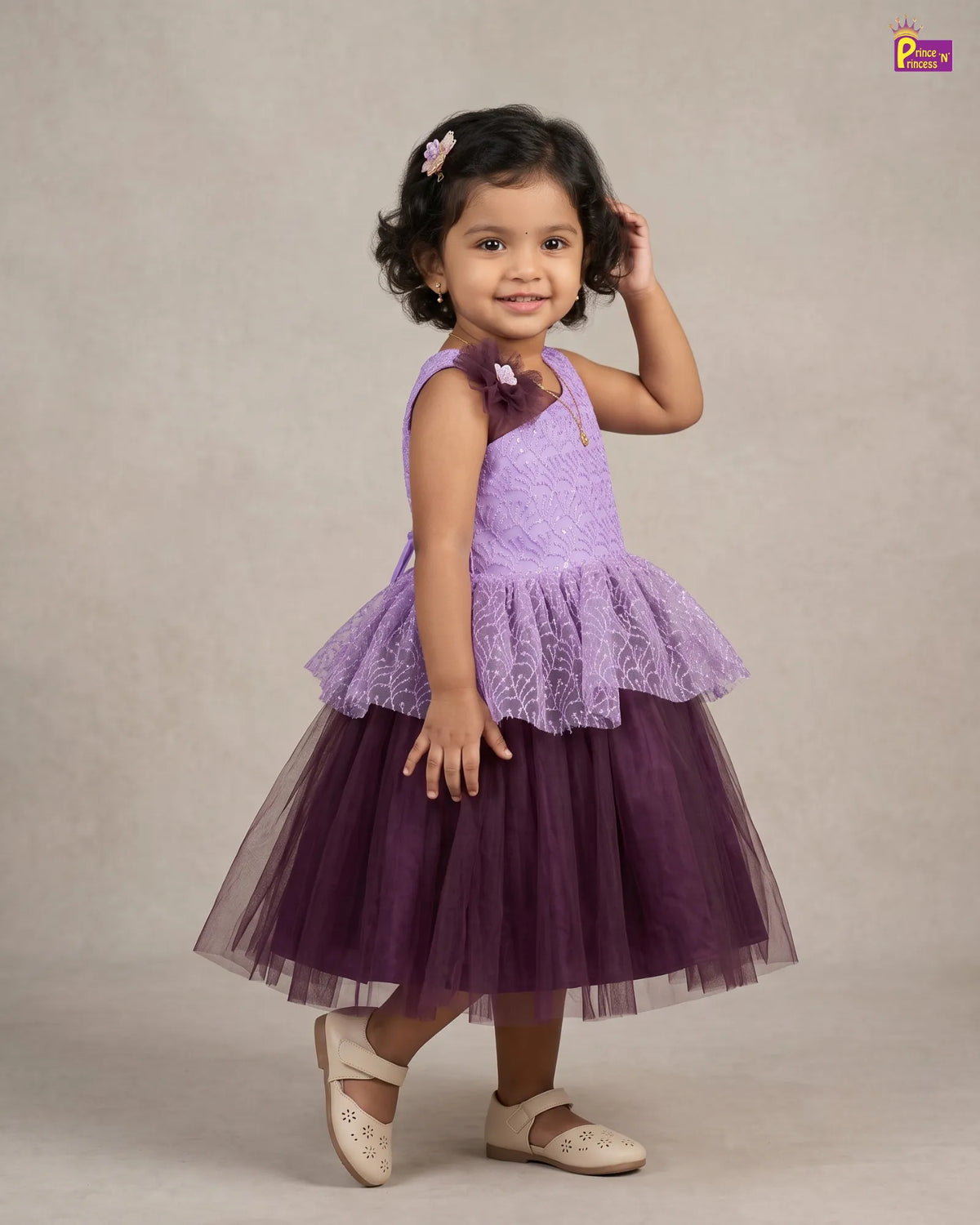 A young girl modeling a two-tone purple dress with a light lavender lace bodice and a darker purple tulle skirt, standing against a neutral beige studio backdrop.
