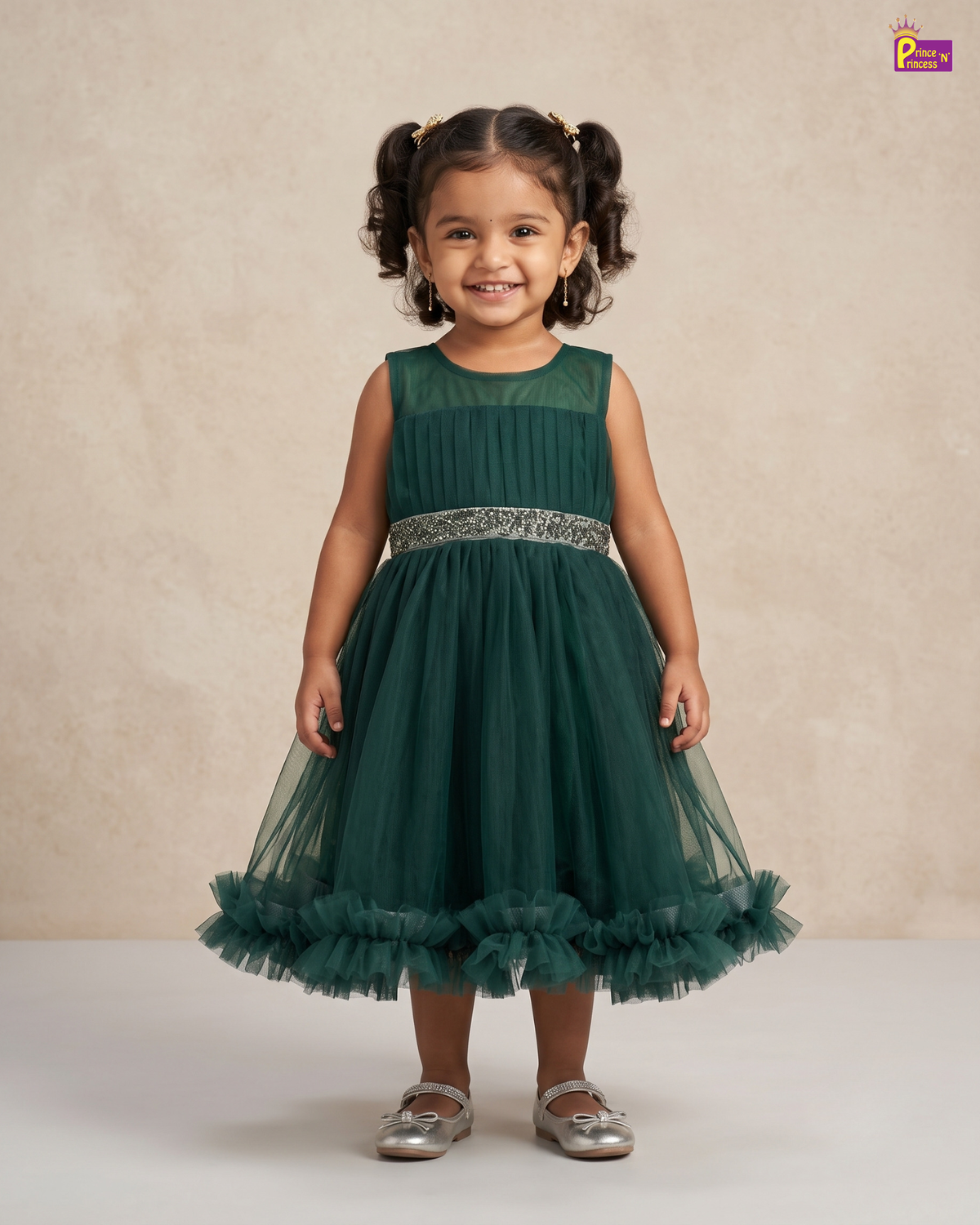 A young girl with dark curly hair in pigtails stands smiling, wearing a sleeveless dark green tulle party dress with a silver beaded waistband and silver shoes, set against a neutral beige textured studio backdrop.