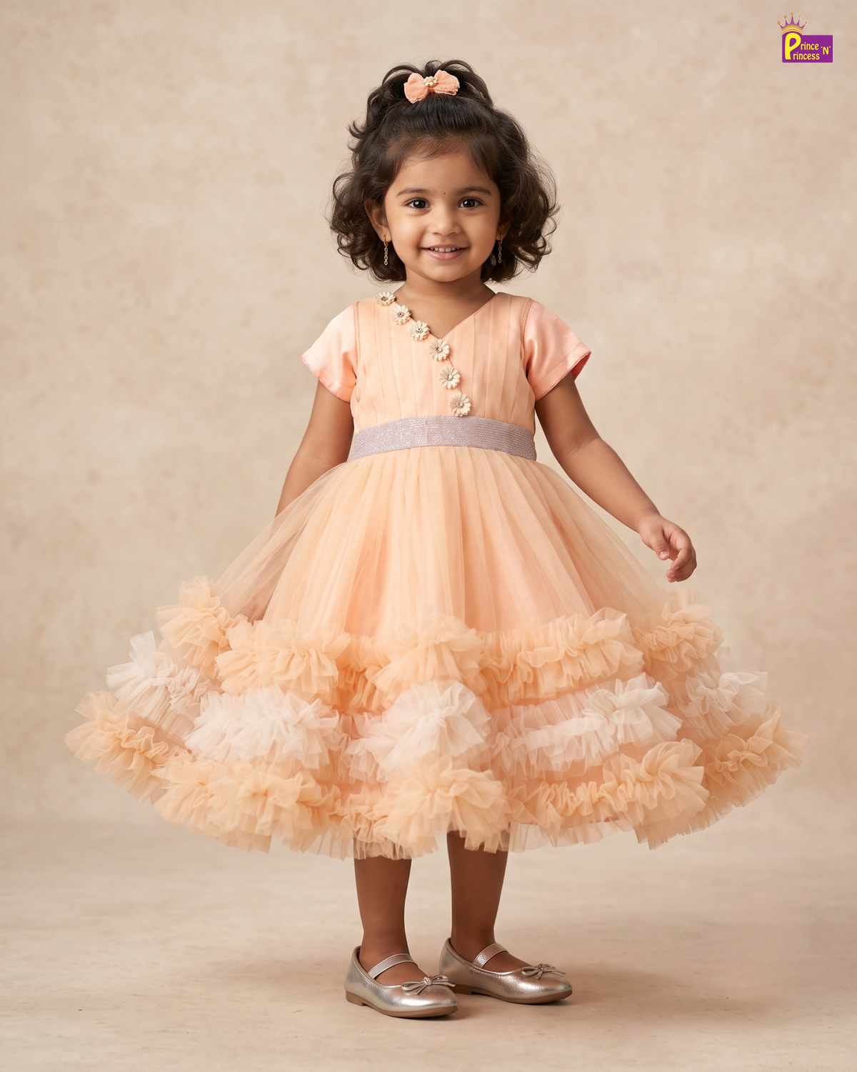 A toddler girl wearing a peach and beige tiered tulle party dress with a lavender waistband and silver shoes, standing against a neutral beige studio backdrop.