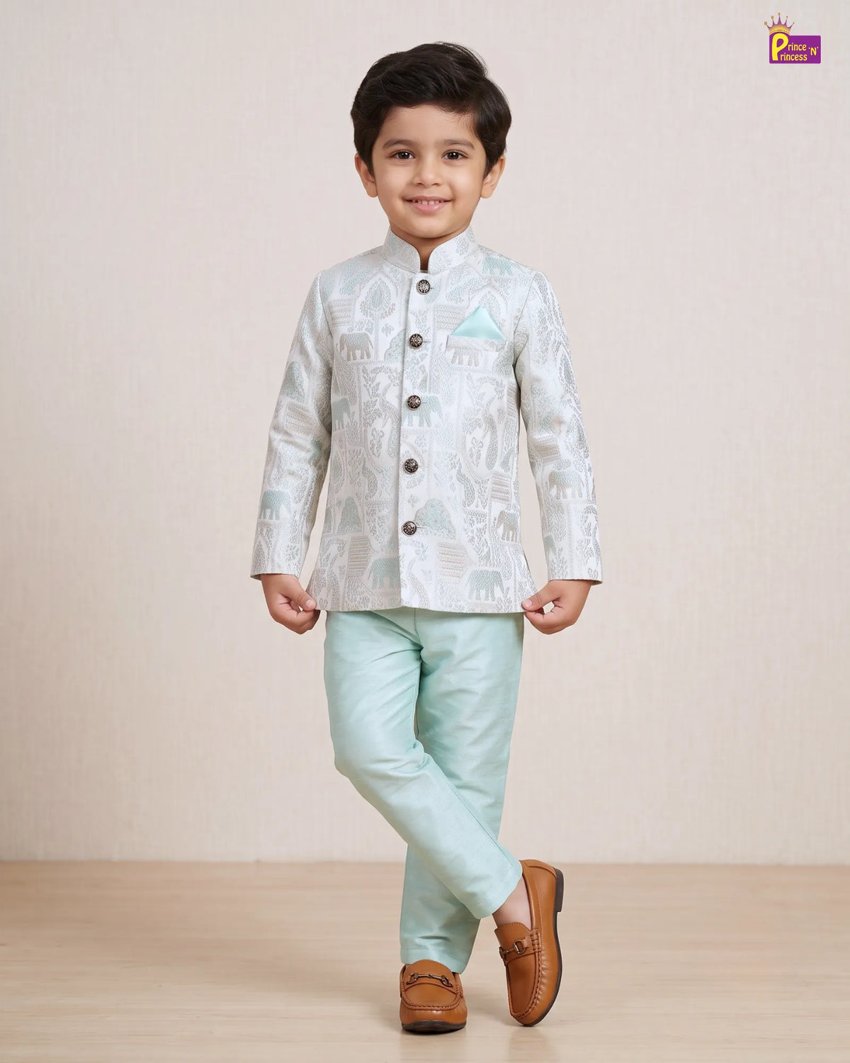 A young boy modeling a traditional Indian outfit consisting of a light silver-gray patterned sherwani jacket with a light blue pocket square, mint green trousers, and brown leather loafers, set against a neutral beige studio background.