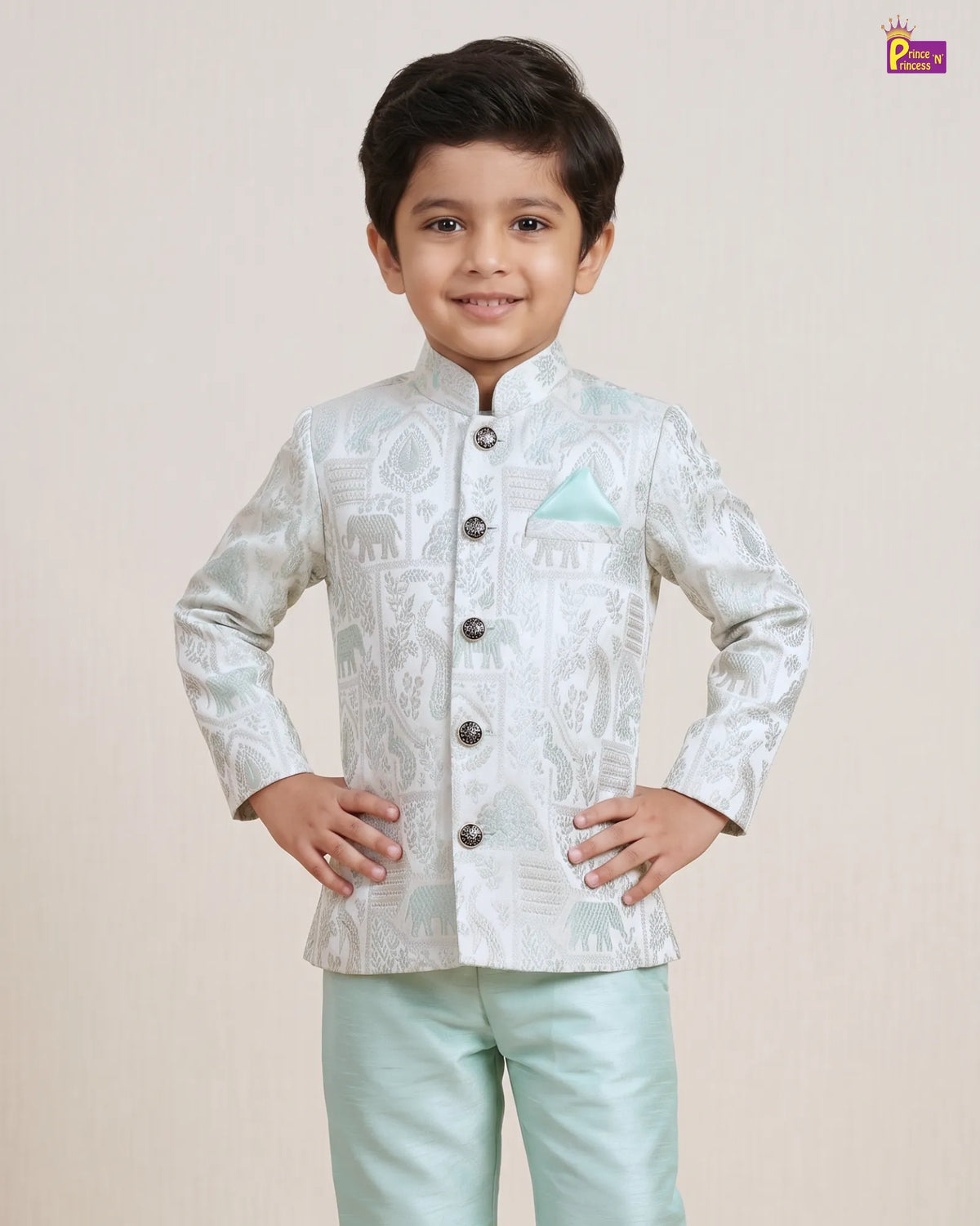 A young boy modeling a light silver-white printed sherwani jacket with a mandarin collar and dark buttons, paired with mint green trousers, against a solid beige background.