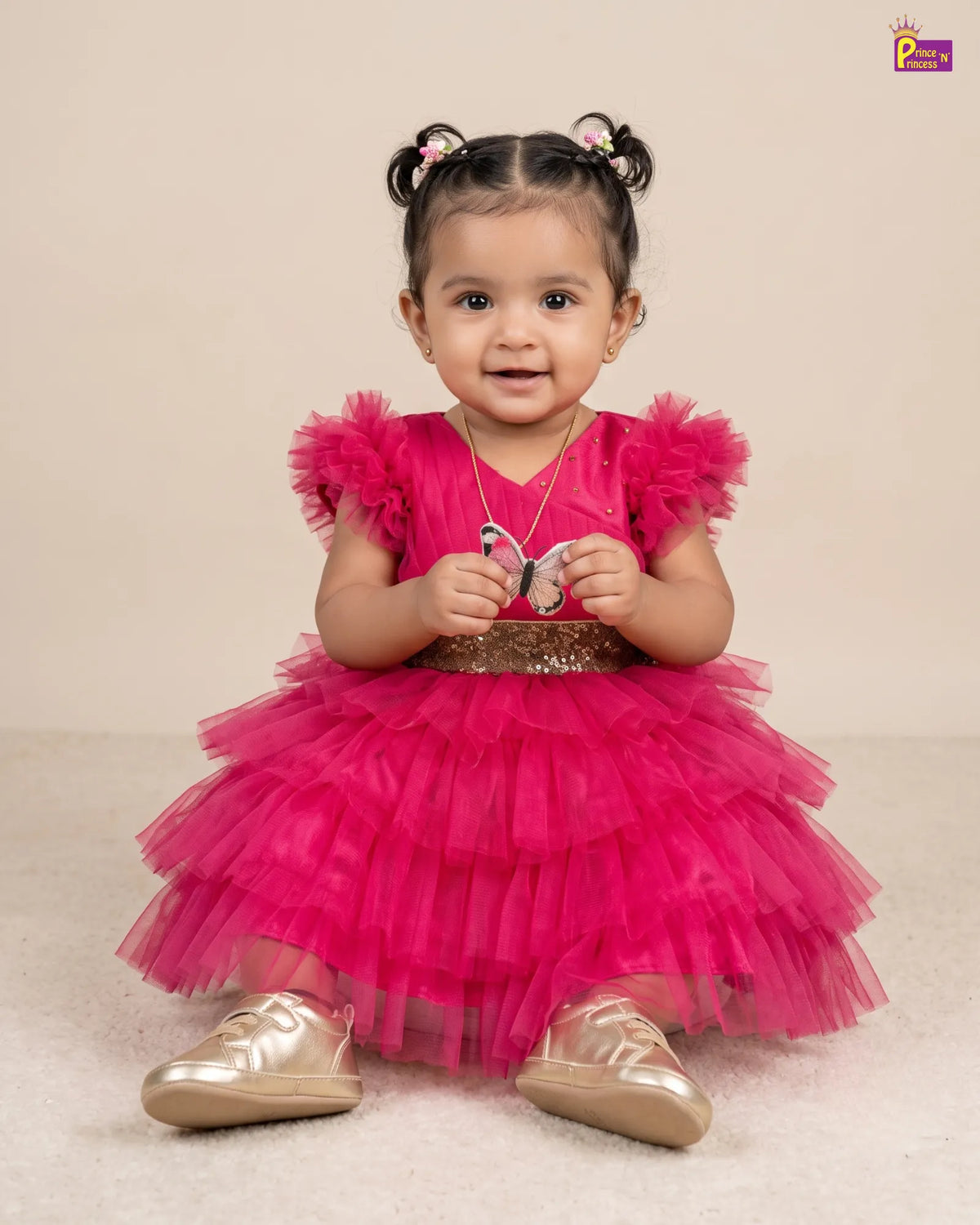 A toddler girl sitting on a beige carpet wearing a bright pink tiered tulle dress with ruffled sleeves and a glittery gold waistband, paired with metallic gold shoes.