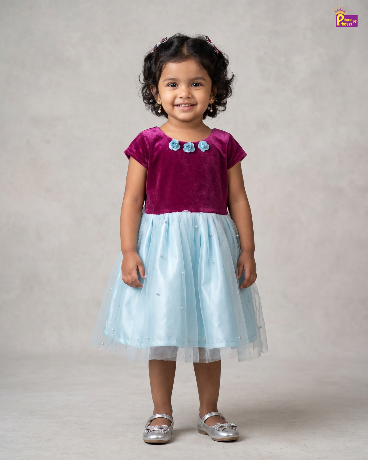 A young girl with curly black hair stands against a neutral beige studio backdrop wearing a two-tone dress with a magenta velvet top and a light blue tulle skirt, paired with silver shoes.