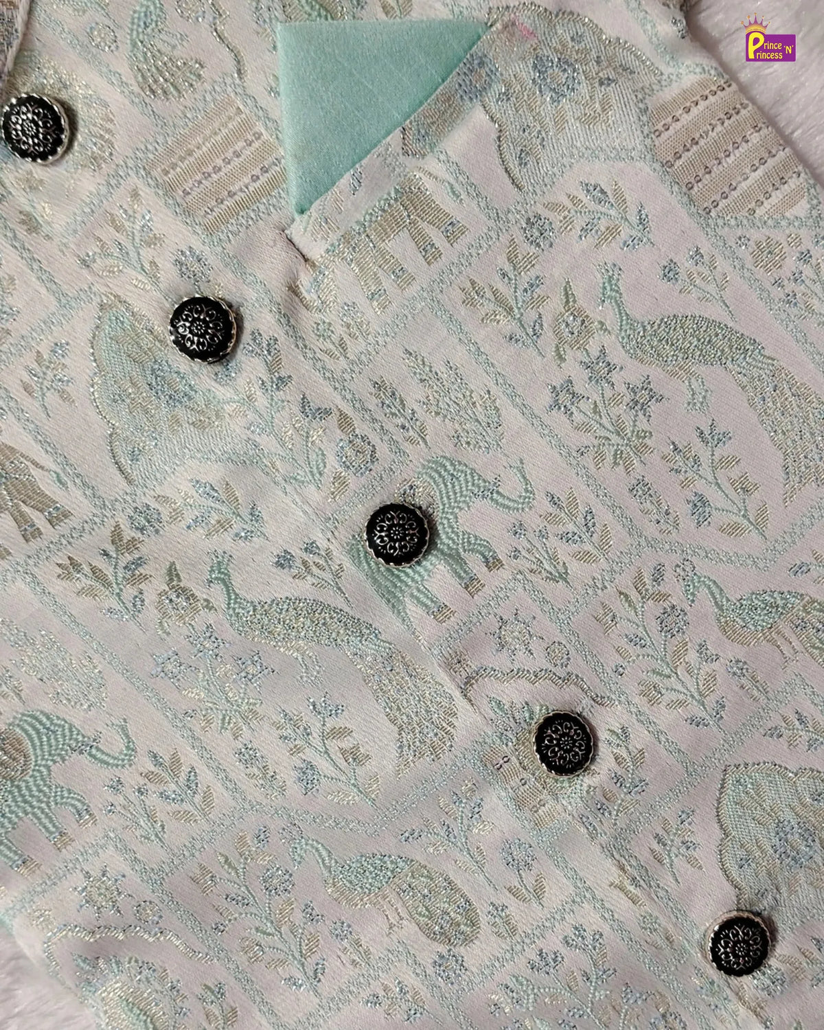 A close-up of a light pink and white embroidered fabric, likely a kurta or shirt, featuring a detailed paisley and floral pattern in pale green and blue, accented with dark metallic buttons.