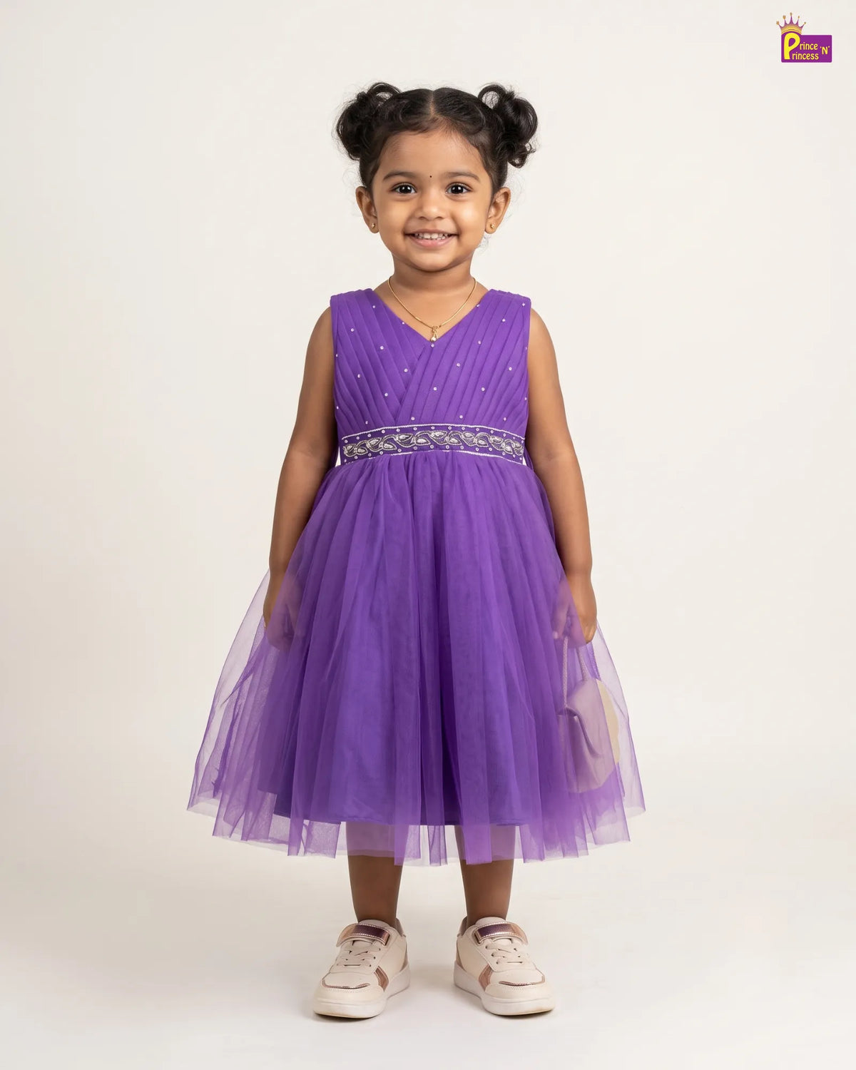 A young girl with her hair in two buns is wearing a sleeveless purple party dress with a tulle skirt and a beaded waistband, standing against a plain off-white background.