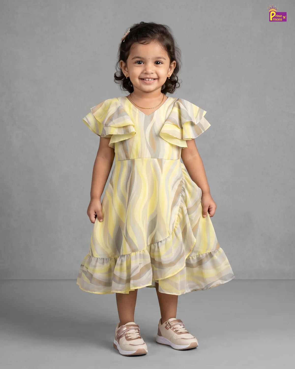 A young girl stands against a gray studio background wearing a yellow and beige wavy-striped ruffled dress with short flutter sleeves and beige sneakers.
