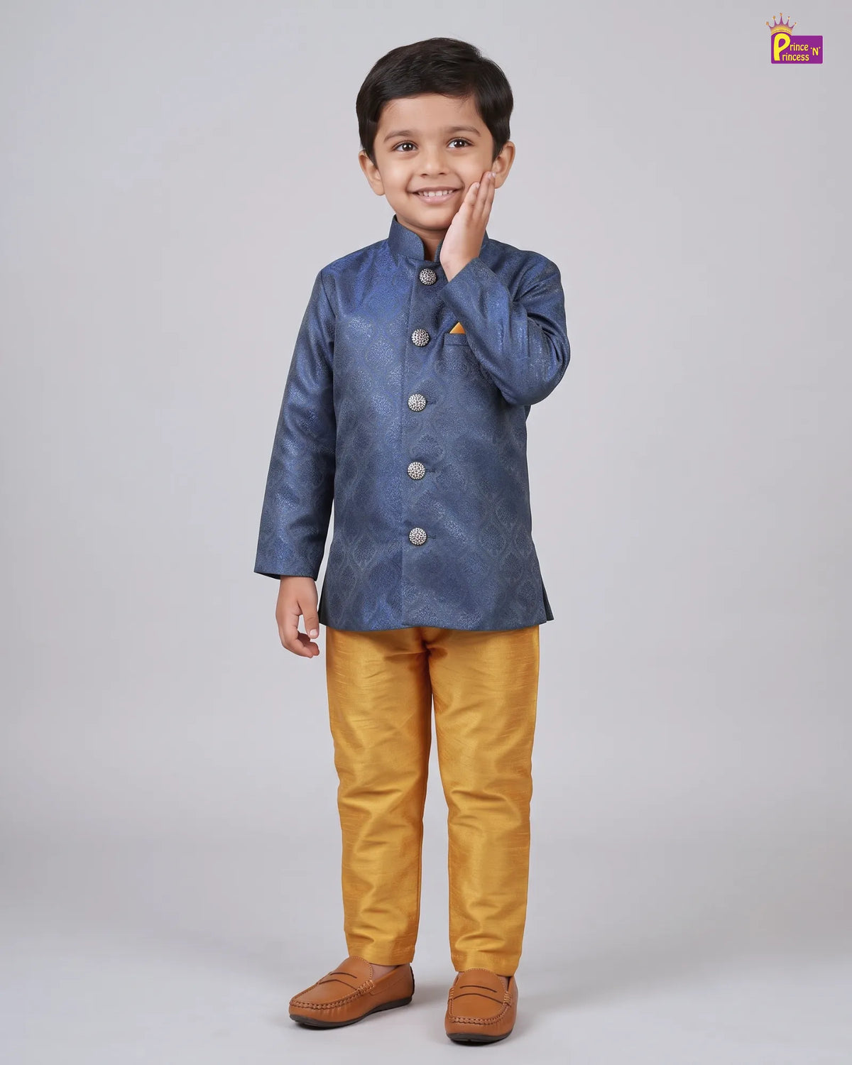 A young boy modeling a traditional Indian outfit consisting of a textured blue long-sleeve jacket and mustard yellow trousers, standing against a plain light gray studio background.