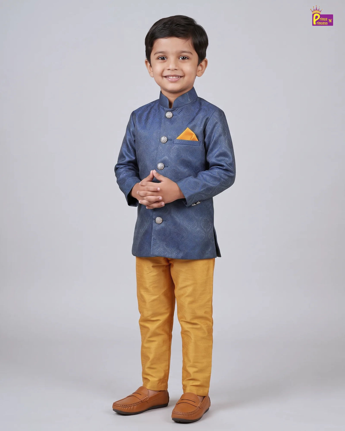 A young boy modeling a traditional Indian outfit consisting of a textured blue long-sleeve jacket with a small orange pocket square, paired with mustard yellow trousers and brown loafers, against a light gray studio background.