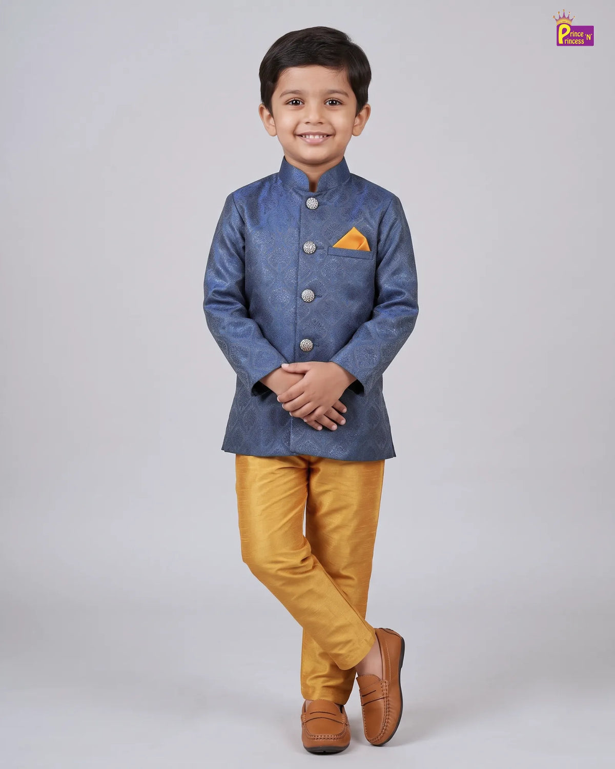 A young boy modeling a traditional Indian outfit consisting of a blue textured sherwani jacket with a yellow pocket square, mustard yellow trousers, and brown leather loafers, set against a plain light gray studio background.