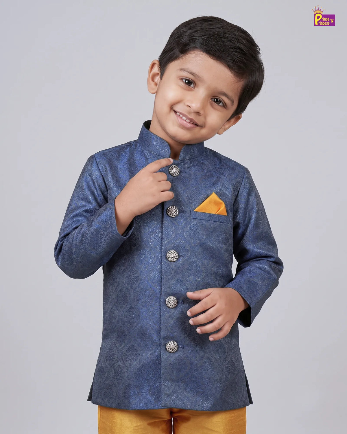 A young boy modeling a blue patterned sherwani jacket with silver buttons and a yellow pocket square, paired with mustard yellow trousers, against a solid gray background.
