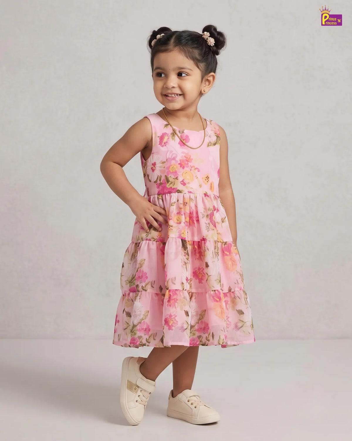 A young girl with her hair in two buns is wearing a pink sleeveless tiered dress with a floral print, standing against a light gray studio background.