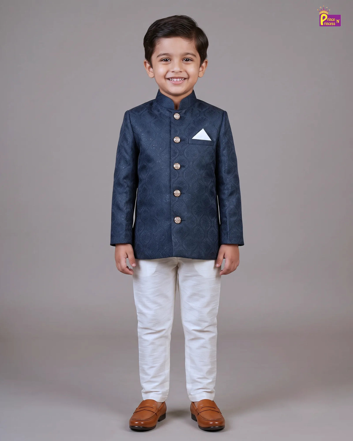 A young boy modeling a dark navy blue textured Indian sherpa jacket with gold buttons, paired with white trousers and brown leather shoes, standing against a neutral gray studio background.