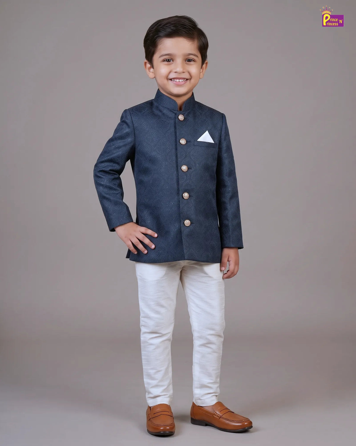 A young boy modeling a navy blue textured sherwani jacket with gold buttons, paired with off-white trousers and brown leather shoes, against a solid gray studio background.