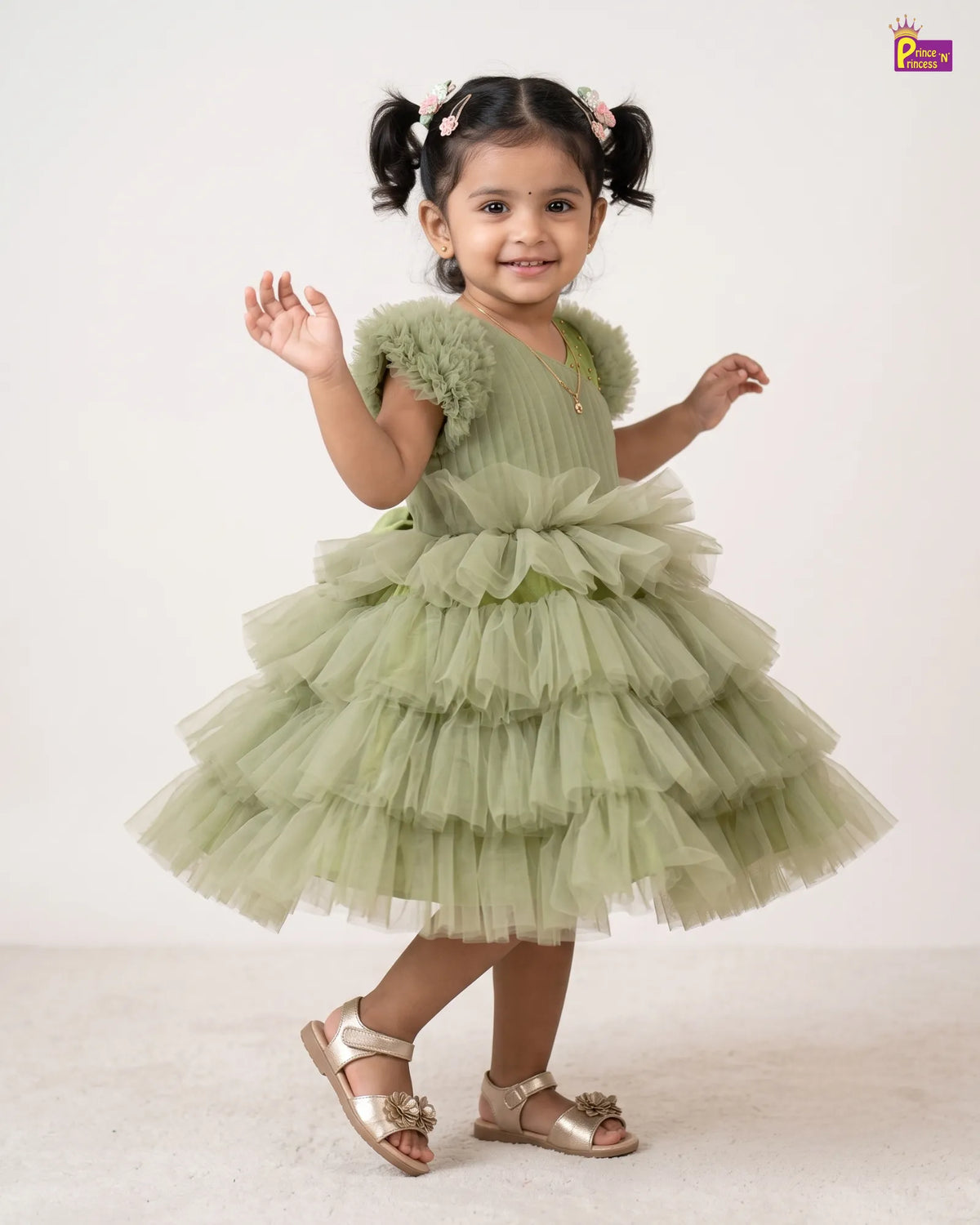 A young girl with dark hair in pigtails models a light sage green tiered tulle party dress with ruffled shoulder details, paired with metallic gold sandals, against a plain off-white studio background.