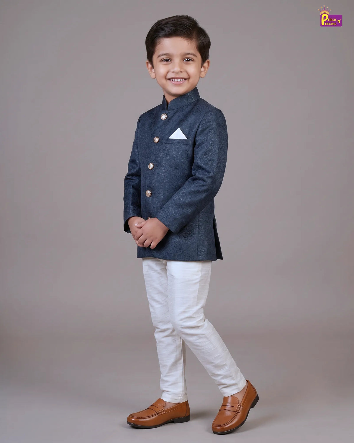 A young boy modeling a navy blue traditional Indian jacket (kurta) with gold buttons, paired with white trousers and brown leather shoes, standing against a solid gray studio background.