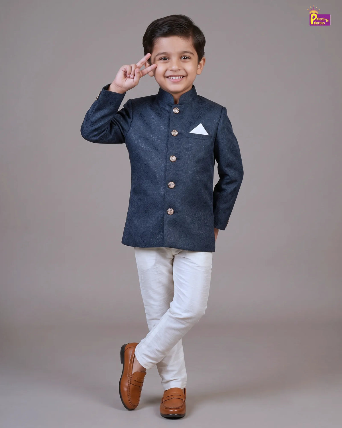 A young boy modeling a dark navy blue textured Nehru jacket with gold buttons and a white pocket square, paired with white trousers and brown leather shoes, posing against a solid gray studio background.