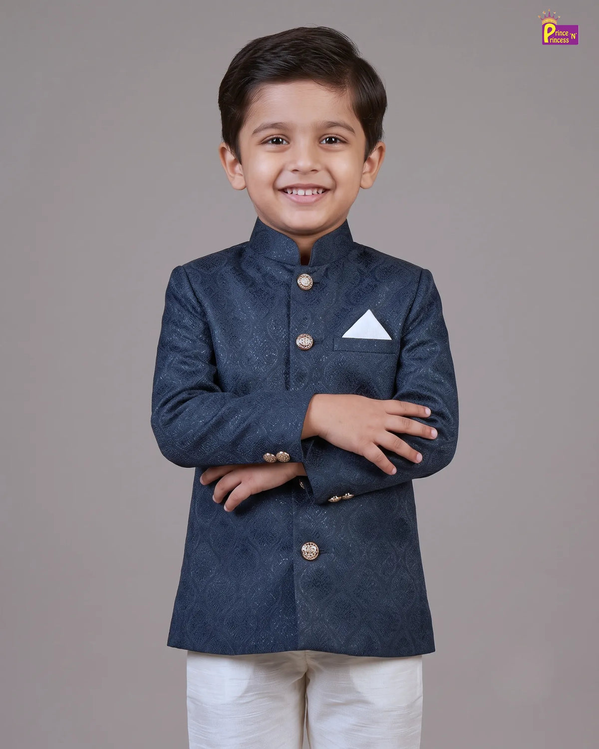 A young boy smiling and posing in a dark navy blue textured sherwani jacket with gold buttons and a white pocket square, paired with white trousers, against a solid gray studio background.