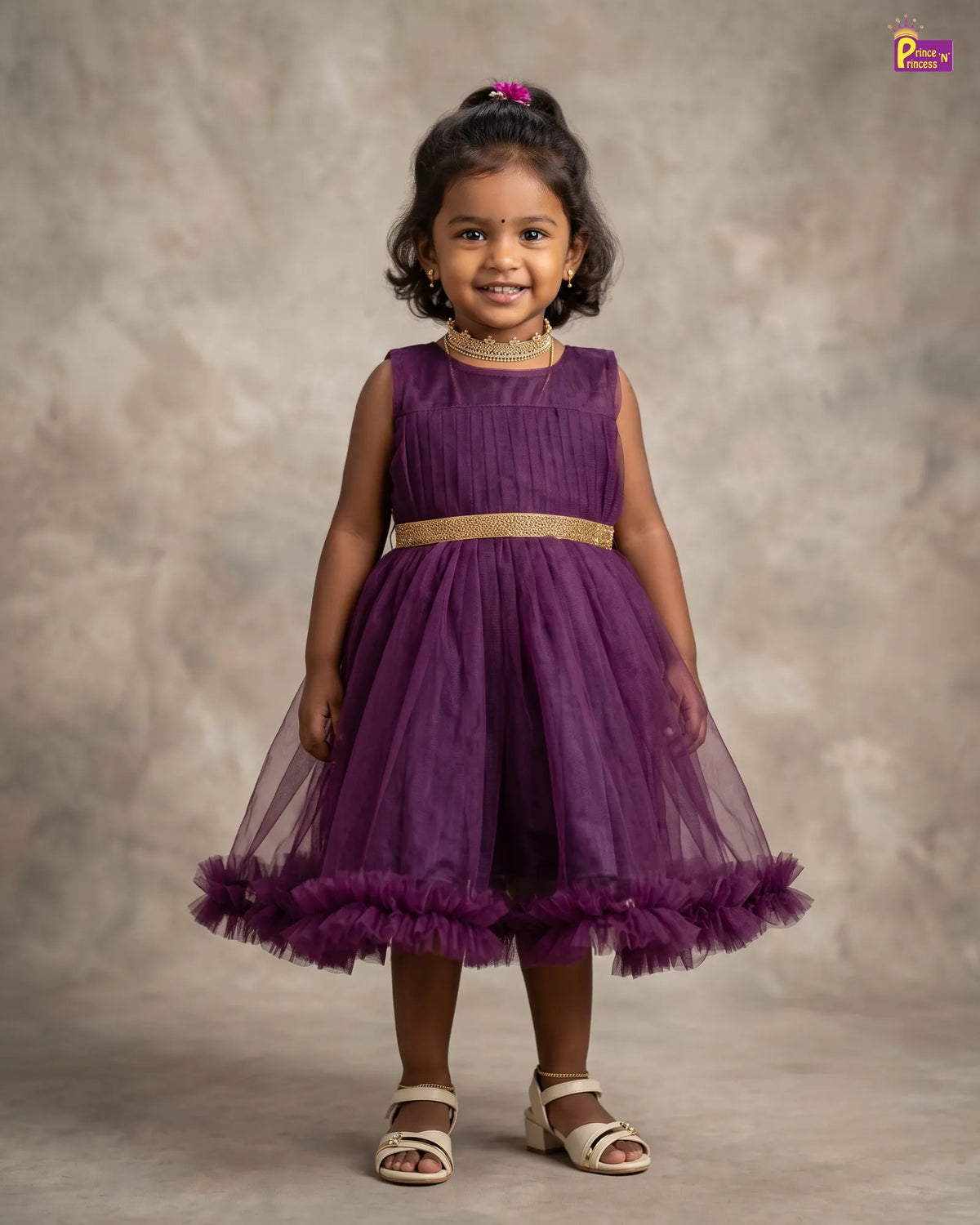 A young girl with dark curly hair wearing a sleeveless purple tulle dress with a gold waistband and beige sandals, standing against a textured gray studio backdrop.