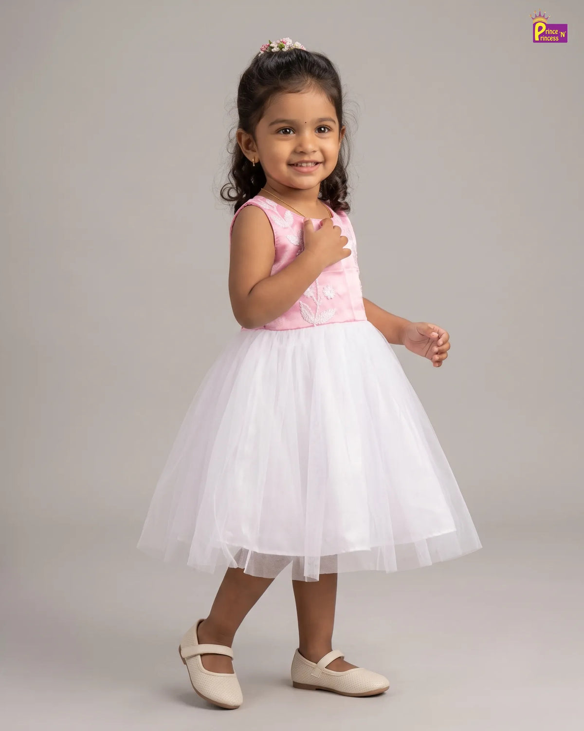 A young girl modeling a sleeveless party dress with a light pink embroidered bodice and a voluminous white tulle skirt, paired with beige ballet flats, against a solid gray studio background.